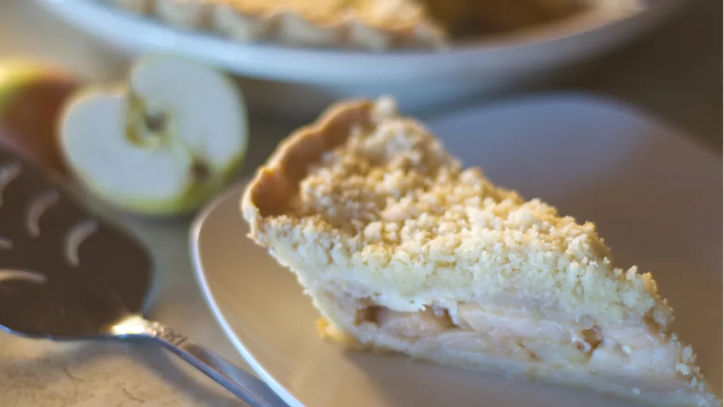White plate with a slice of Apple Crumb Pie on it. In the background is the rest of the pie and a halved apple.