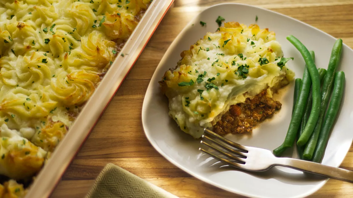 A plate with a serving of shepherd's pie topped with parsley, accompanied by green beans. A fork rests on the plate. Next to it, there is a baking dish filled with more shepherd's pie. The setting is on a wooden table with a cloth napkin.