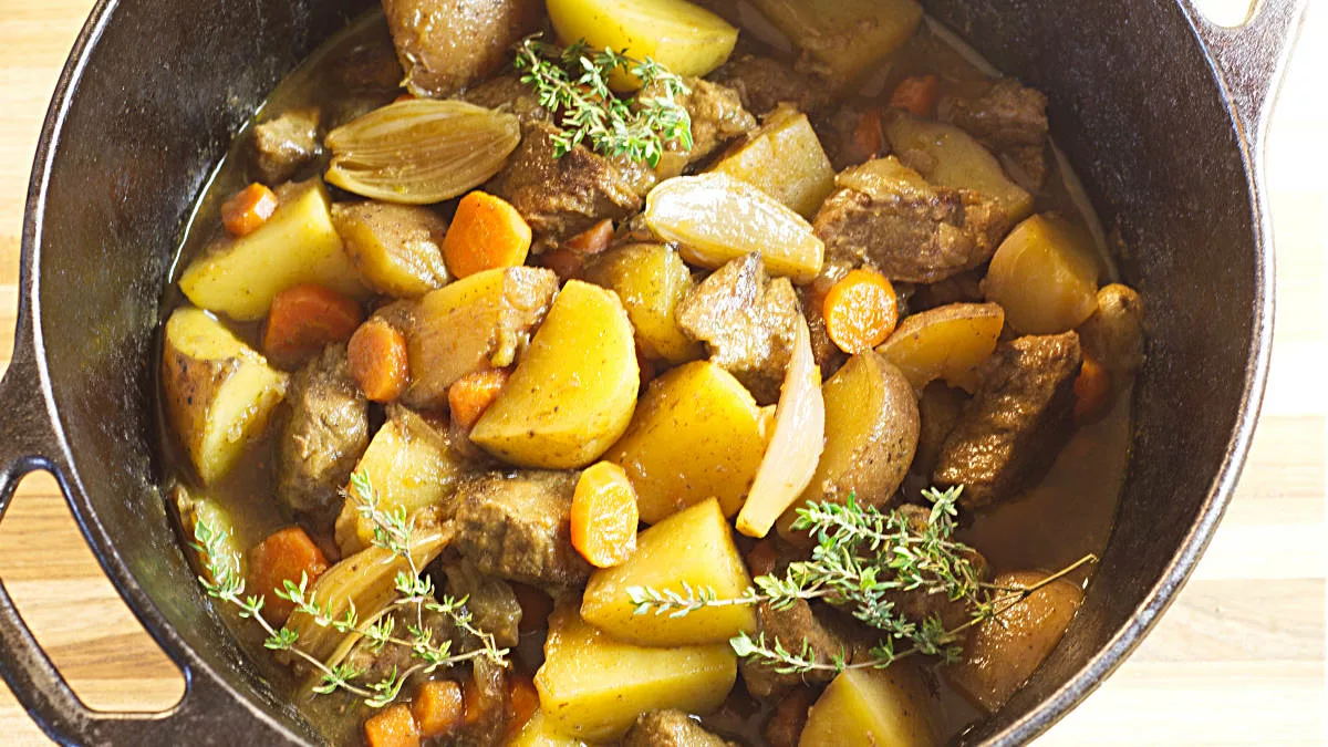 A hearty beef stew in a black pot, filled with chunks of beef, potatoes, carrots, and onion slices. The dish is garnished with several sprigs of fresh thyme. The stew appears to be rich and flavorful, with a thick broth coating the ingredients.
