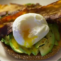 Poached egg laying on sliced Avocado with bacon on a toasted half sesame bagel.