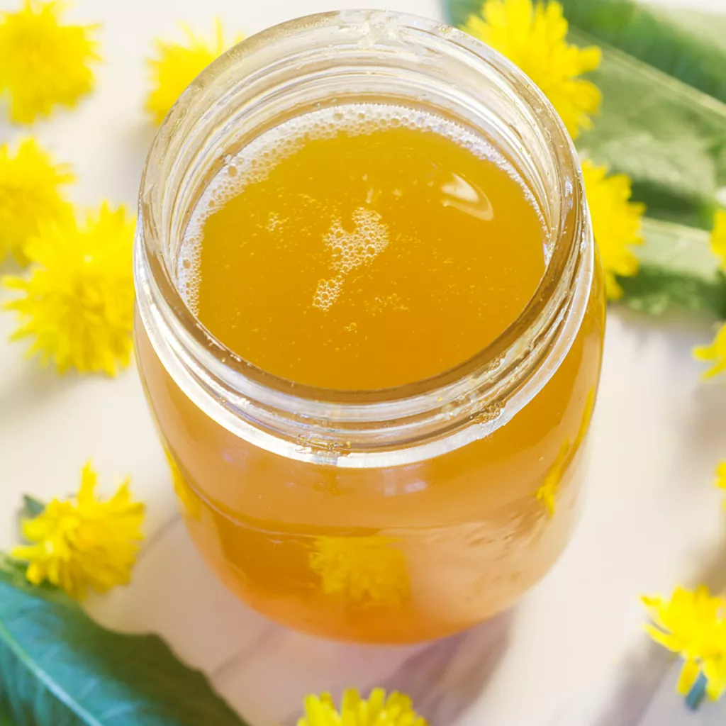 Jar of Dandelion Jelly with Dandelion flowers and leaves laying around.
