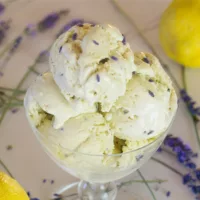 Glass dish containing multiple scoops of Lemon Lavender Ice Cream, garnished with some fresh lavender buds, with lemons and lavender in the background.