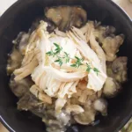 Dark bowl containing stuffing, and mushrooms topped with shredded chicken and garnished with a sprig of thyme.
