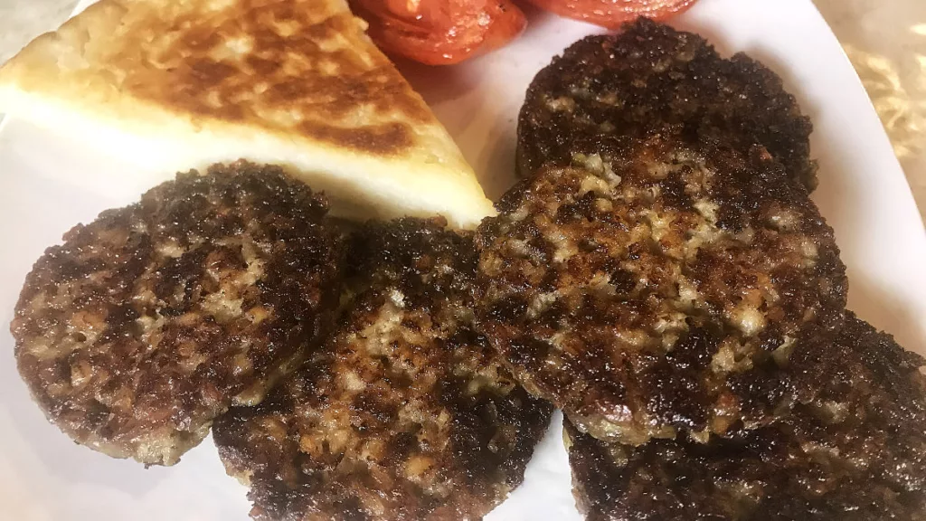 A white plate containing 5 slices of Black Pudding with potato bread and tomato in the background.