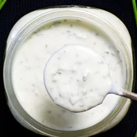 Spoon coming out of a jar of Crème Fraîche Dressing. Jar is on a black background.