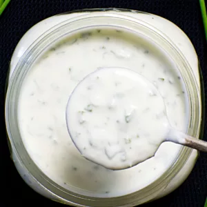 Spoon coming out of a jar of Crème Fraîche Dressing. Jar is on a black background.