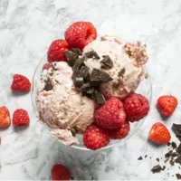 Glass dish containing Raspberry Chocolate Ice Cream garnished with chocolate chunks and fresh raspberries.