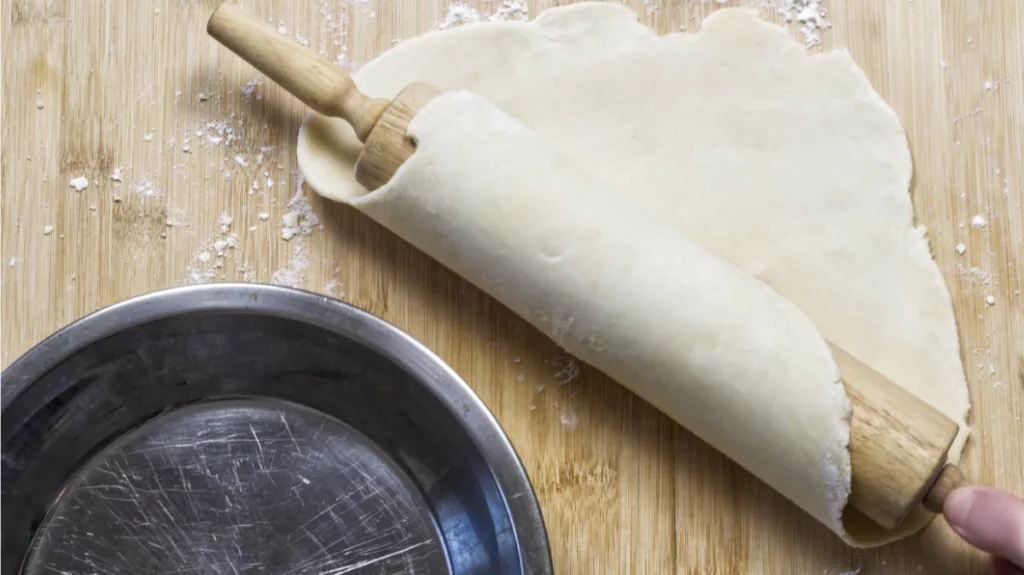 Rolled out pastry being lifted by a rolling pin. Pie dish is on the side.
