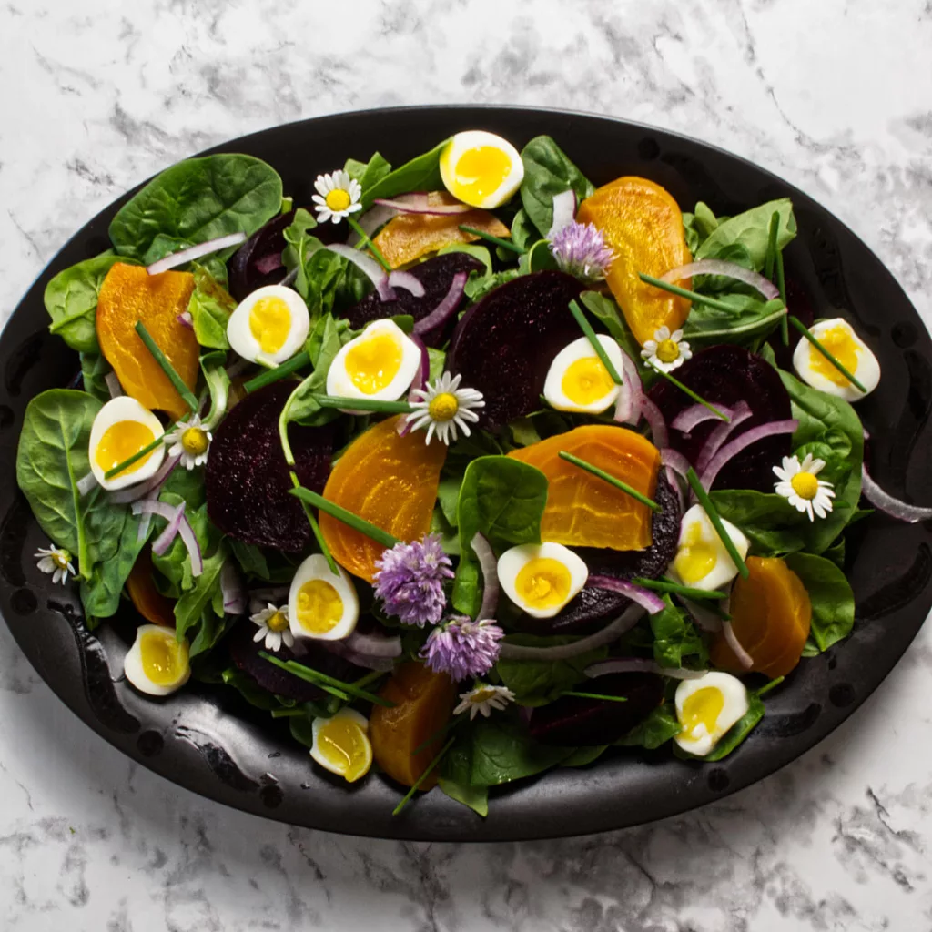 Black oval platter showing Spinach and Arugula salad with quail eggs, roasted beets, red onion, garnished with chives and chamomile flowers.