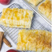 Wire rack containing two Apple Cinnamon Pop Tarts. Another two tarts are in the background on a baking sheet. Fresh apples and cinnamon sticks used as garnish.