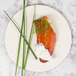 White plate containing a filet of Hot Smoked Salmon garnished with a Crème Fraîche Dressing and dill and fresh chives.