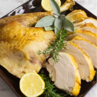 Dark square platter containing sliced roasted whole turkey from frozen. Garnished with fresh rosemary, sage and lemon.