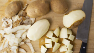 Wooden board with potatoes on it. Some are chopped, some peeled and some unpeeled. A chopping knife is off to the side.