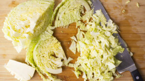 Wooden board with Savoy cabbage on it, some of it is shredded the rest is in tack. Chopping knife is resting on the board.
