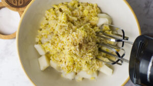 Cabbage and onion mixture added to the bowl of boiled potatoes, salt and pepper added and a hand held mixer ready to combine everything.