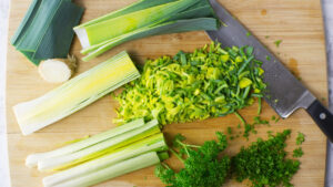 Wooden board containing washed and chopped leeks, parsley and a chopping knife.