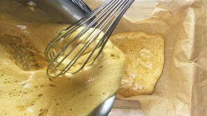Yellowman batter being poured from a pot, using a whisk, into a container lined with parchment paper