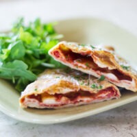 Pepperoni Pizza Calzone that has been cut to show the filling on green plate. Also on the plate is a side of arugula lettuce.