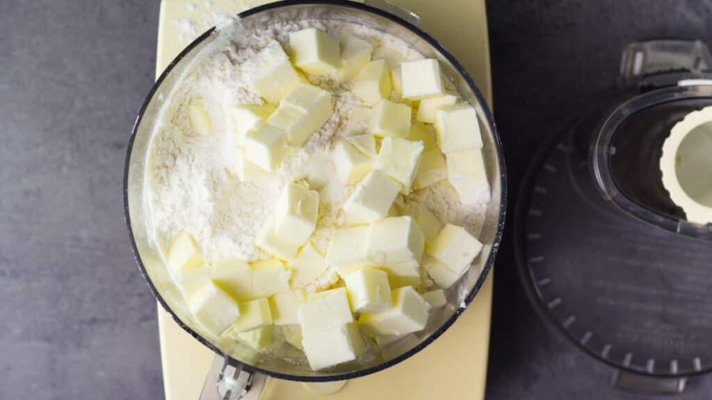 Food processor bowl filled with cubed up butter and flour. To the side is the lid of the processor.