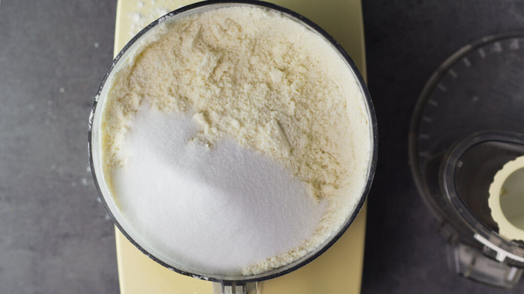 Food processor bowl filled with the mixed up butter and flour. Sugar is added to the mixture. To the side is the lid of the processor.