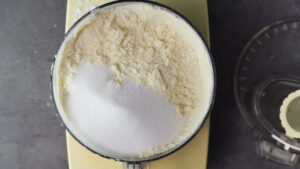Food processor bowl filled with the mixed up butter and flour. Sugar is added to the mixture. To the side is the lid of the processor.