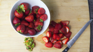 Wooden board with a white bowl containing strawberries on it. On the board is also some strawberry tops, and some sliced strawberries and a chopping knife.