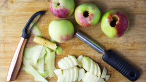 Wooden board with apples on it. Some of the apples have been cored, some peeled and sliced. The is also a peeler and corer resting on the board.