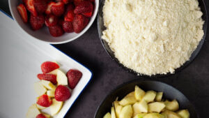 A white baking dish containing some apples and strawberries is on the left with a bowl of strawberries beside it. Also beside it is a bowl of crumb mix and a bowl of sliced apples. This is the assembling of the Apple and Strawberry Crumble.