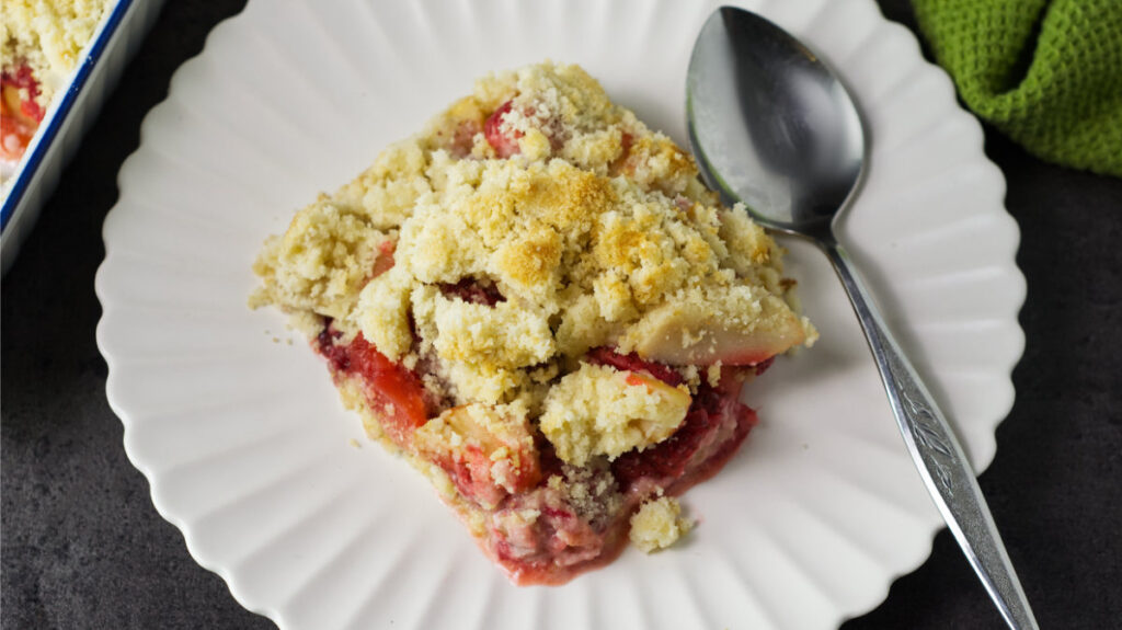 A round, white plate with a piece of Apple and Strawberry Crumble on it. A spoon is resting on the plate, in the background is a dish containing the rest of the Apple and Strawberry Crumble and on the right is a dish towel.