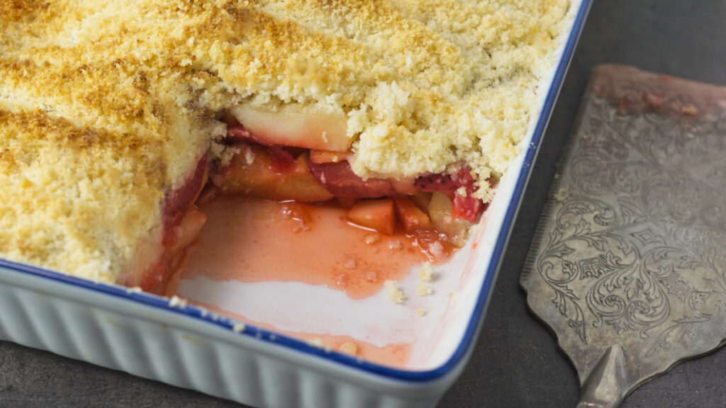 White baking dish containing the Apple and Strawberry Crumble with a piece removed from it. On the side is a serving utensil.