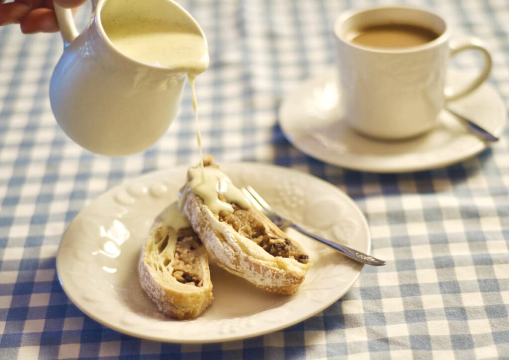White page with 2 slices of Apfelstrudel on it. Vanilla sauce is being poured on it. In the background is a cup of coffee. The tablecloth is blue and white checkered.