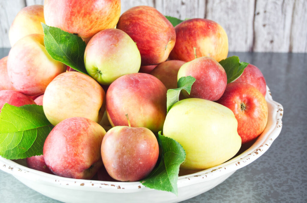 A large, cream bowl of apples with apple leaves dispersed throughout the apples.