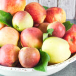 A large, cream bowl of apples with apple leaves dispersed throughout the apples.