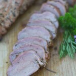 Wooden board with 2 Pork Tenderloins. One loin is sliced. Garnishing the board are fresh herbs and chive flowers.
