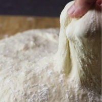 A wooden board with Slow Rise Pizza dough on it being pulled showing dough elasticity.