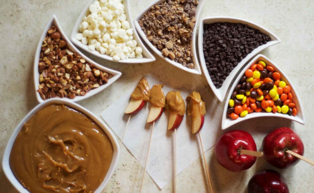 Candy Apple Dipping Bar showing 5 toppings, a bowl of caramel, slice apples that have been dipped in the caramel and some whole apples with sticks in them.
