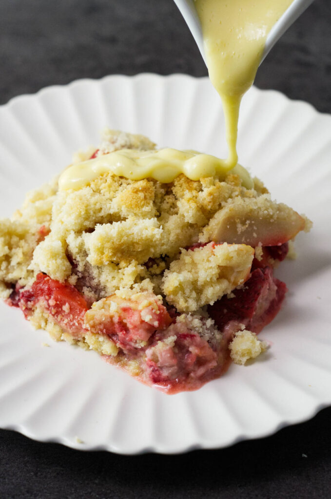 A white plate containing a portion of Apple and Strawberry Crumble with yellow custard being poured onto it.