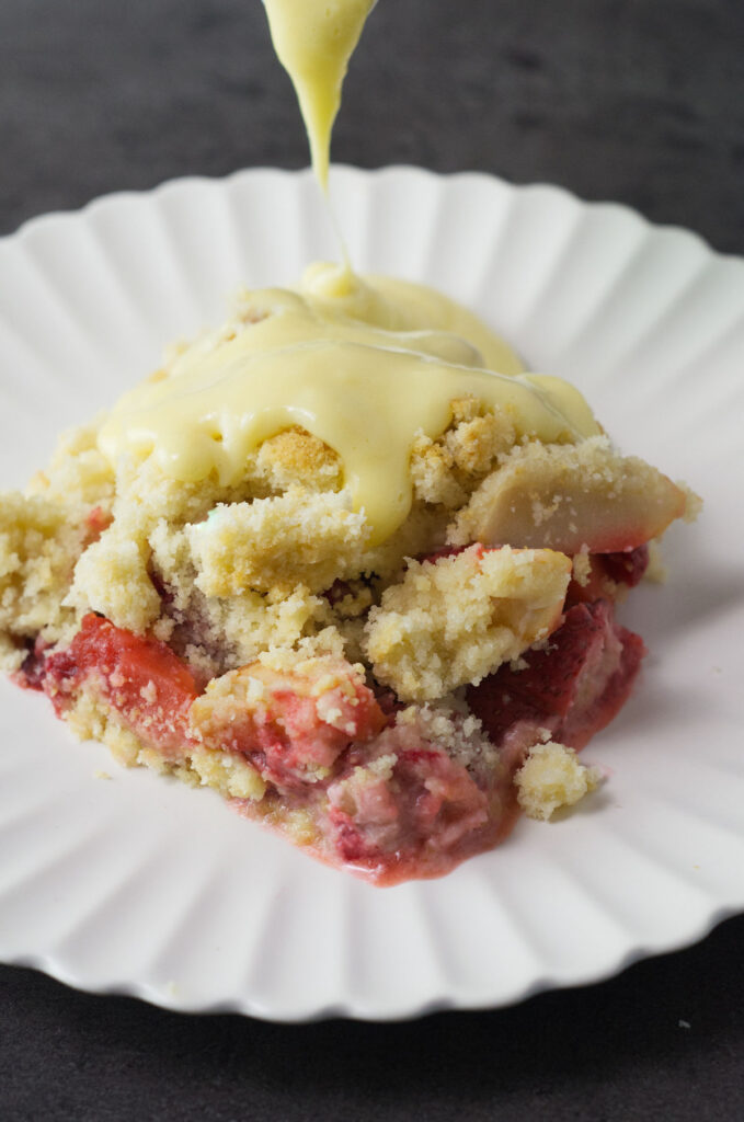 A white plate containing a portion of Apple and Strawberry Crumble with yellow custard being poured onto it.