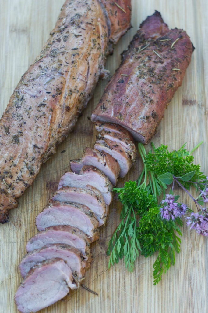 Wooden board with 2 Pork Tenderloins. One loin is sliced. Garnishing the board are fresh herbs and chive flowers.
