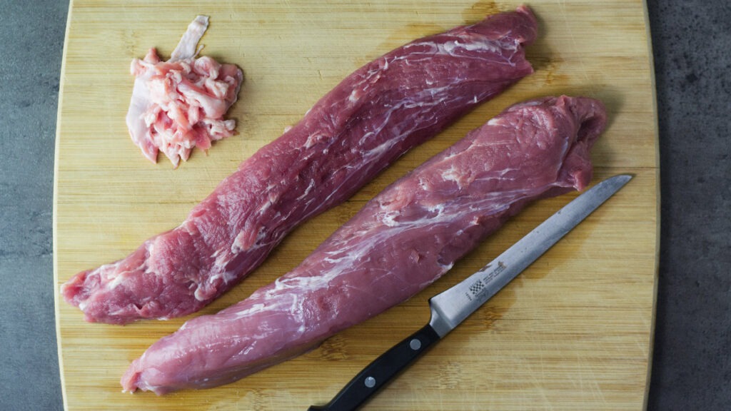 Wooden board with two raw pork tenderloins on it, trimmed away fat and silver skin are sitting on the board, also the knife is on the board.