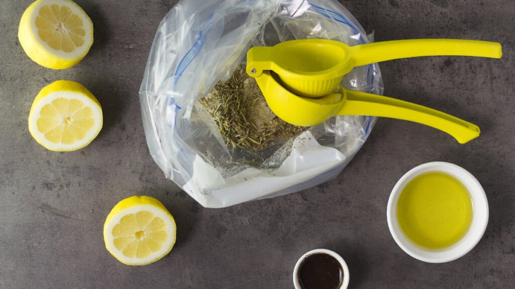 Fresh lemon juice is being squeezed into the ziplock bag. Surrounding the bag are halved lemons, olive oil and Worcestershire sauce.