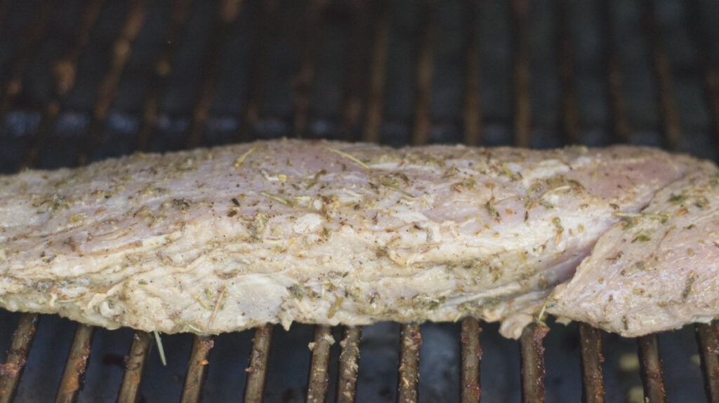 Raw pork tenderloin on a grill grate. The tenderloin is covered in herbs.