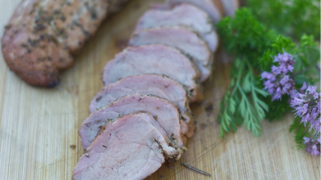 Wooden board with Pork Tenderloins. One loin is sliced. Garnishing the board are fresh herbs and chive flowers.