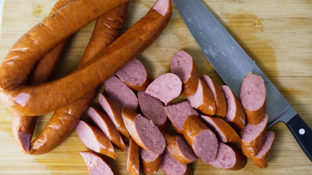 On a chopping board are some whole sausages along with some sliced ones and a chopping knife is resting on the board.