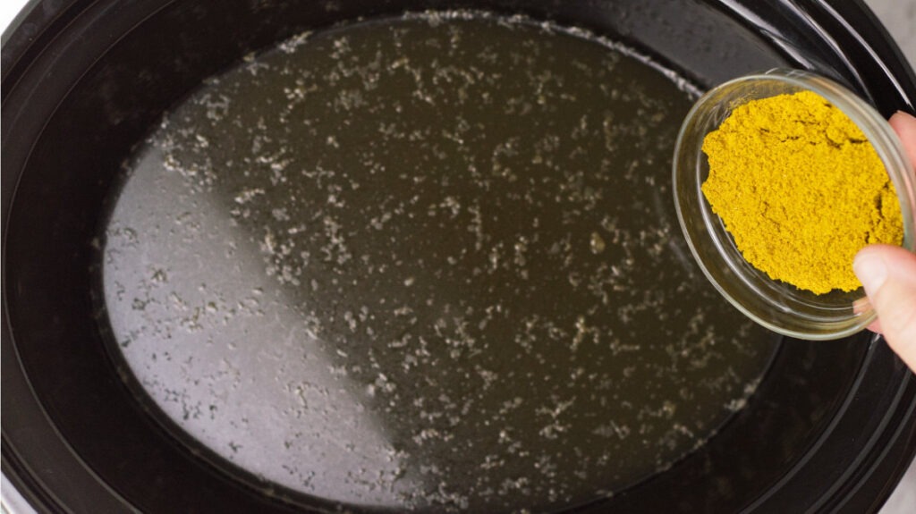 Slow cooker insert containing stock with a small bowl of curry powder about to be added. 