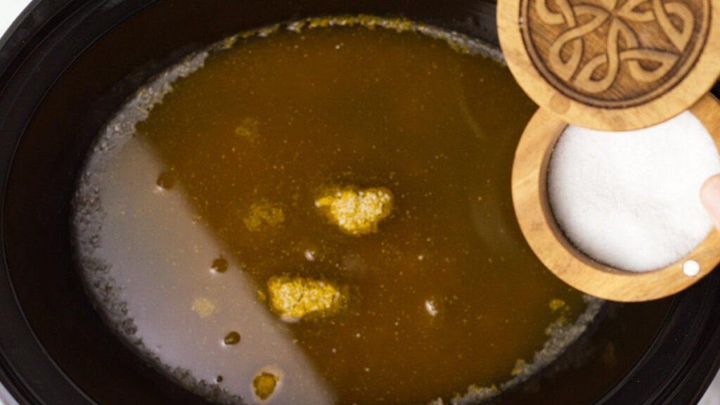 Slow cooker insert containing stock with a salt container at the side, inferring that salt will be added to the stock.