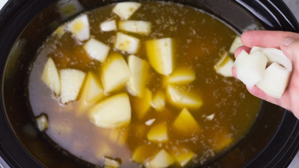 Potatoes and onions are added to the stock in the slow cooker. To the right is a hand showing some potatoes that have yet to be added.
