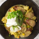 Up close image of a black bowl containing slow cooker curried sausages garnished with a dollop of Greek yogurt, fresh herbs and a flowering pea vine.