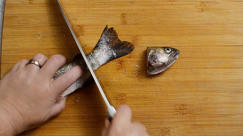 Wooden board with a rainbow trout on it, its head has been removed and the tail is being cut off. One hand is holding the fish and the other hand is holding a knife used to remove the tail.