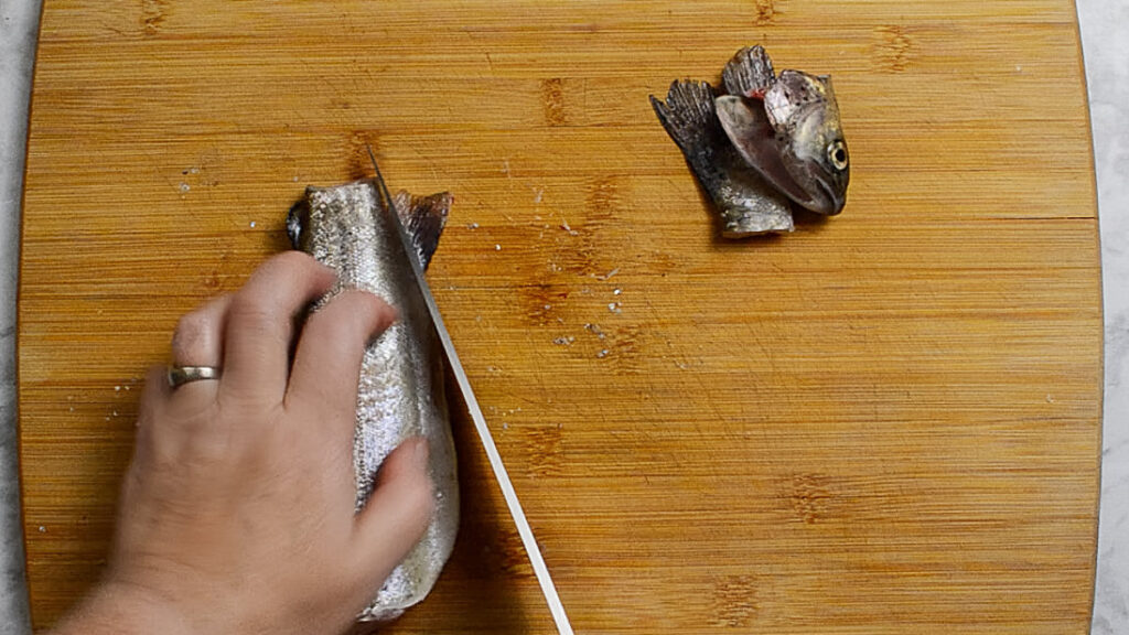 Wooden board with a rainbow trout on it, the head and tail are on the right of the board, and on the left is the body of the fish with the fins being sliced off. A hand is holding the fish and a knife is slicing through the fins.